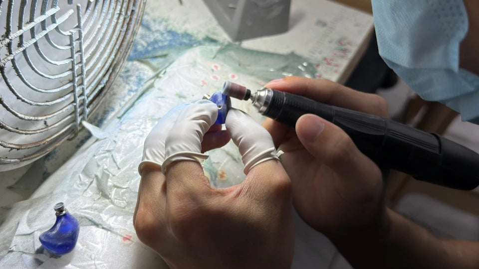 A close-up of a jeweler's hands wearing white gloves, using a rotary diamond grinding tool to shape a small blue gemstone, with a metal fan and workbench visible in the background.