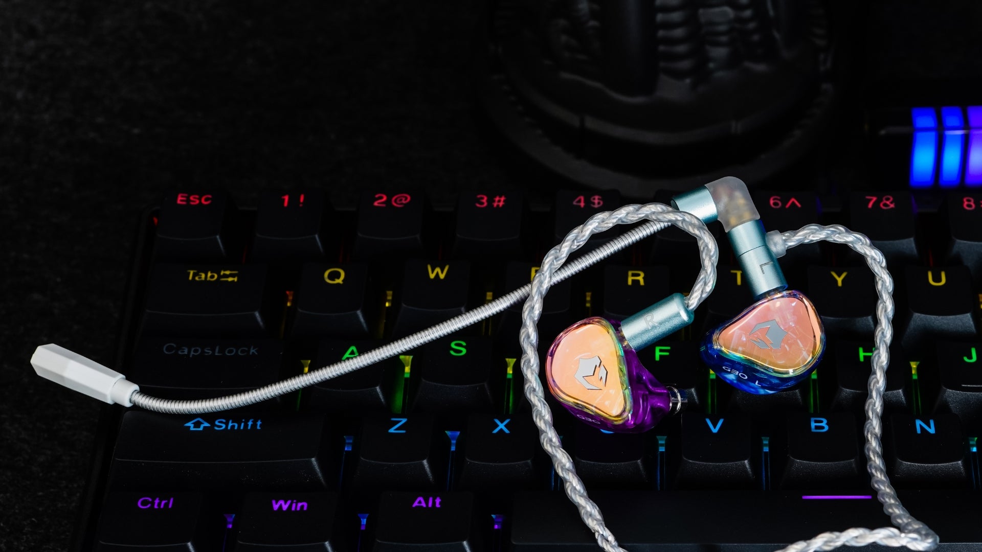A pair of high-end in-ear monitors with a silver braided cable, resting on a black mechanical keyboard with RGB backlighting.