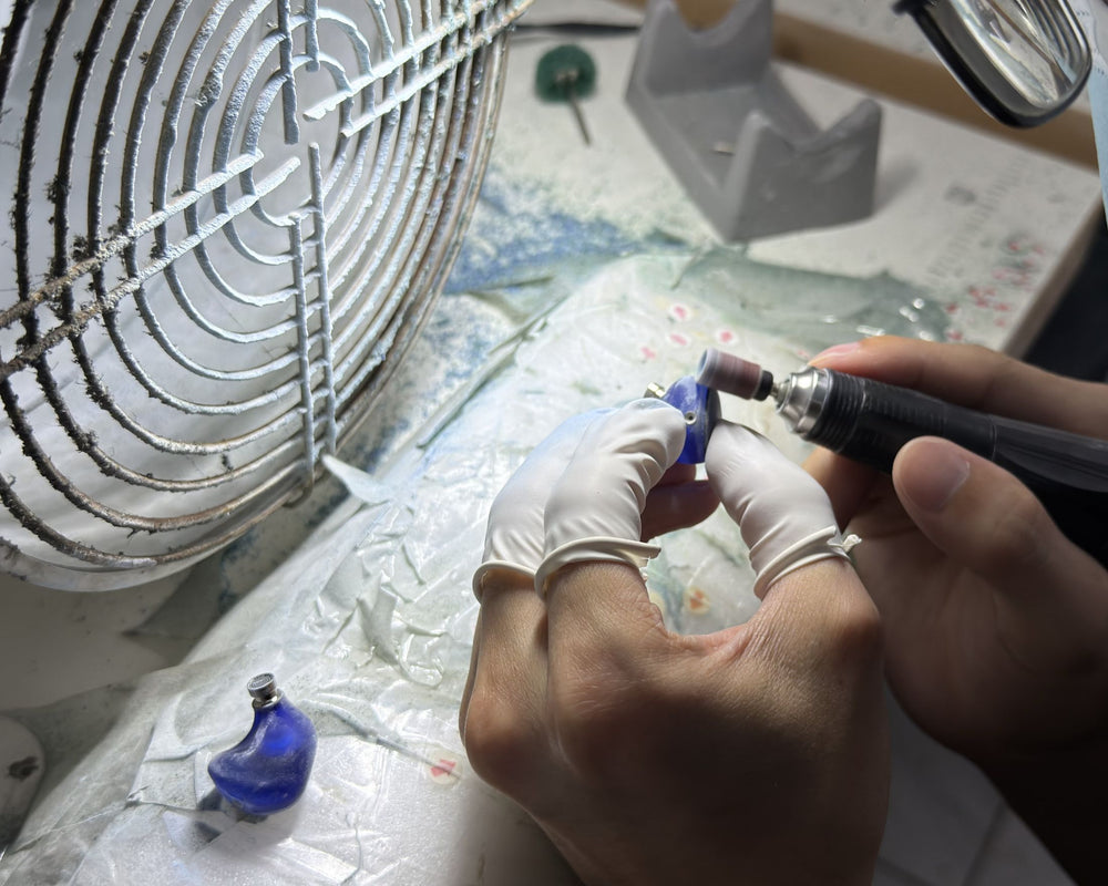 A close-up of a jeweler's hands wearing white gloves, using a rotary burr with a sanding attachment to work on a small blue gemstone or jewel setting. A metal fan is visible in the background, along with a light source and workbench materials.