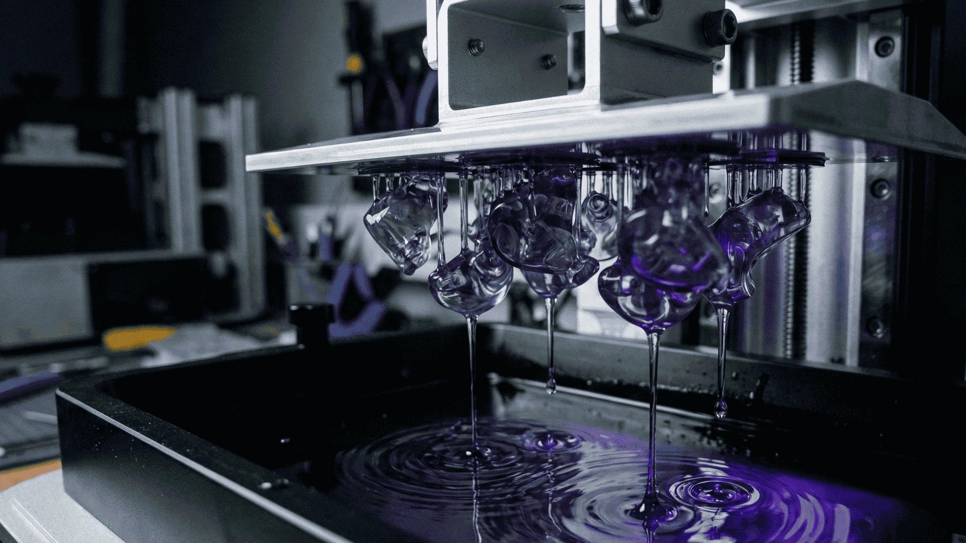 A close-up of a 3D printer's print head depositing purple transparent filament in a tray, with the filament dripping in long strands and forming circular ripples on the surface.